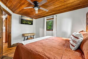 A bedroom with a wooden ceiling, a bed with a patterned bedspread, a nightstand, a desk, a window with a view of trees, and a painting on the wall. - NEW Dolly Fireworks View! 3 King, LuxHotTub, Fence Yd (Pigeon Forge)