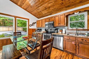 A kitchen with wooden cabinets, stainless steel appliances, and a glass dining table with chairs. - NEW Dolly Fireworks View! 3 King, LuxHotTub, Fence Yd (Pigeon Forge)