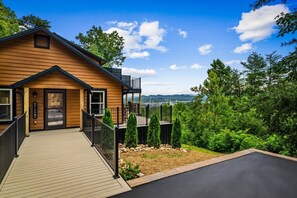 A wooden cabin with a deck, a balcony, and a view of a forested landscape. - NEW Dolly Fireworks View! 3 King, LuxHotTub, Fence Yd (Pigeon Forge)