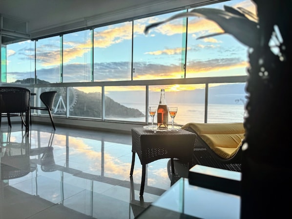 A room with a large window overlooking a sunset, a bottle, and two glasses on a table.