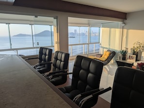 Una habitación moderna con una mesa grande y sillas, un sofá, y vista al mar y al horizonte de la ciudad.