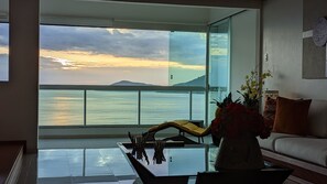 Ein modernes Wohnzimmer mit einem großen Glasfenster, das auf einen Balkon mit Blick auf ein Gewässer und eine ferne Berglandschaft führt.