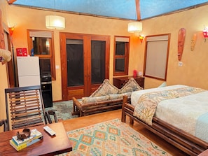Una habitación con una cama, un banco de madera, una mesa con libros, un refrigerador y una ventana.