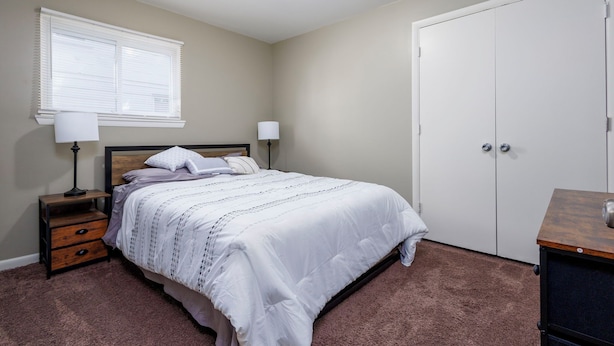 A bedroom with a bed, two bedside tables, a window with blinds, a door, and a carpeted floor. - 3/1 Home in Southfield, Near Hospitals & Parks (Southfield)