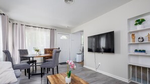 A modern living room with a flat-screen TV, a dining area with a table and chairs, and a small bookshelf with decorative items. - 3/1 Home in Southfield, Near Hospitals & Parks (Southfield)