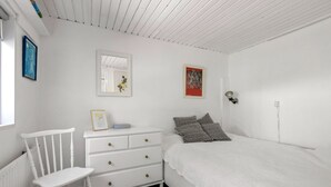 Et soveværelse med en hvid seng, en kommode, en stol og en vægmonteret lampe.