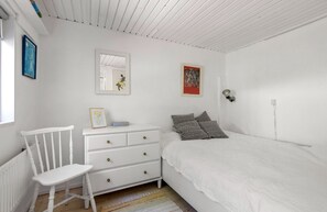 A bedroom with a white bed, a dresser, a chair, and a wall-mounted lamp. - Idyllic holiday home with large garden (Fårevejle)