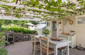 Une terrasse couverte avec une table et des chaises, un banc en bois, un pot suspendu et un espace jardin agrémenté de plantes et d’une treille.