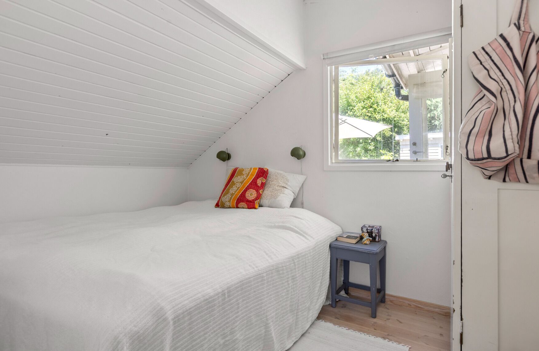A bedroom with a sloped ceiling, a bed with a white bedspread, a small bedside table with a lamp, and a window with a view of greenery outside.