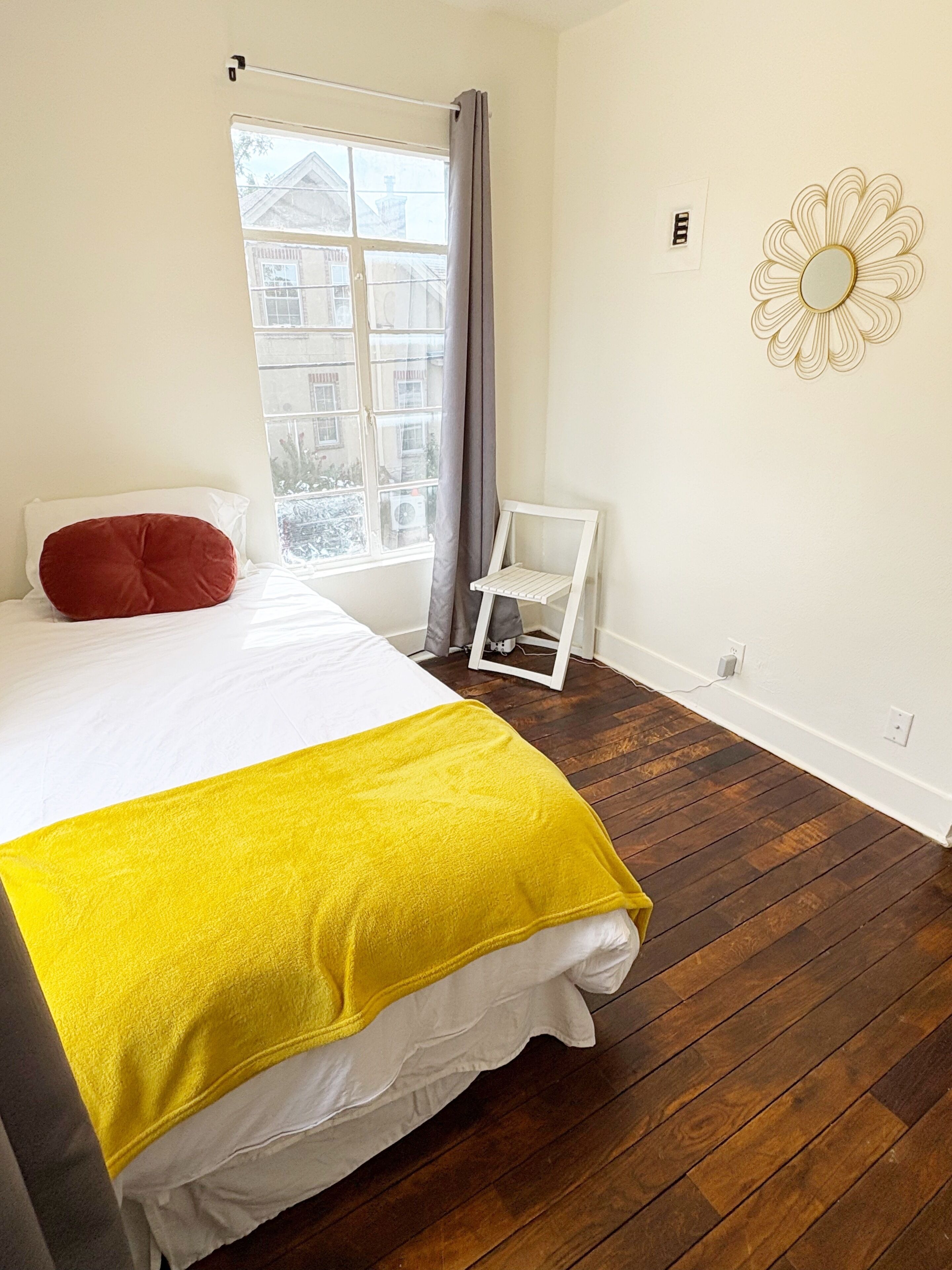A bedroom with a bed, a window with curtains, a chair, and a wall-mounted mirror.
