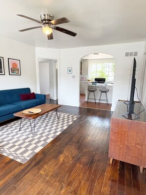 A living room with a blue sofa, a wooden coffee table, a checkered rug, a ceiling fan, and a kitchen area in the background.