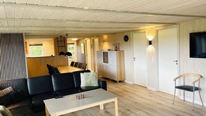 Un intérieur moderne avec un plafond en bois, un coin repas comprenant une longue table et des chaises, un canapé noir, une petite table avec deux tasses et un téléviseur fixé au mur.