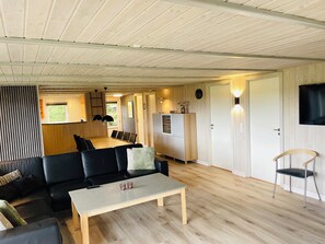 A modern interior with a wooden ceiling, a dining area with a long table and chairs, a black sofa, a small table with two cups, and a TV mounted on the wall. - Spacious holiday home in West Jutland (Nørre Nebel)