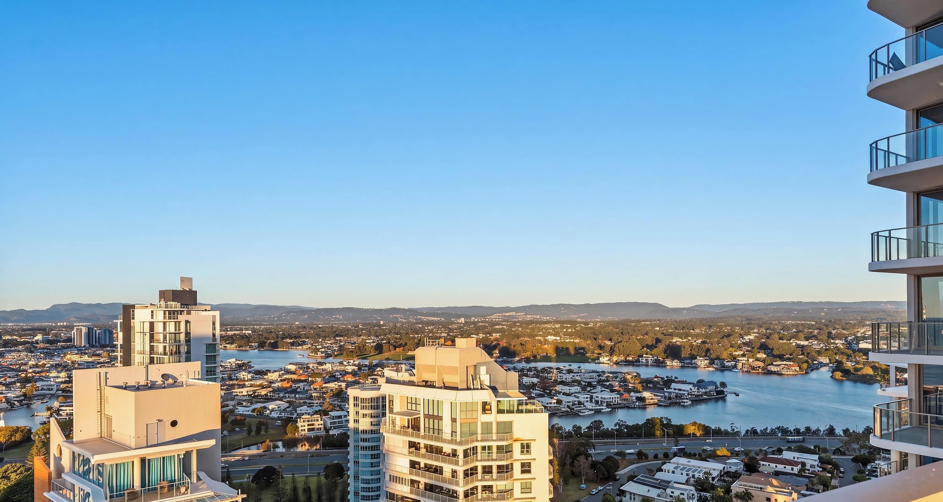 A high-rise building with multiple balconies overlooking a coastal cityscape with a river and distant hills.