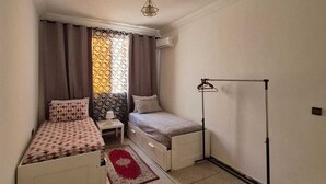 A small, compact bedroom with two beds, a bedside table, a floor lamp, and a red rug. - Confort plus (Agadir)