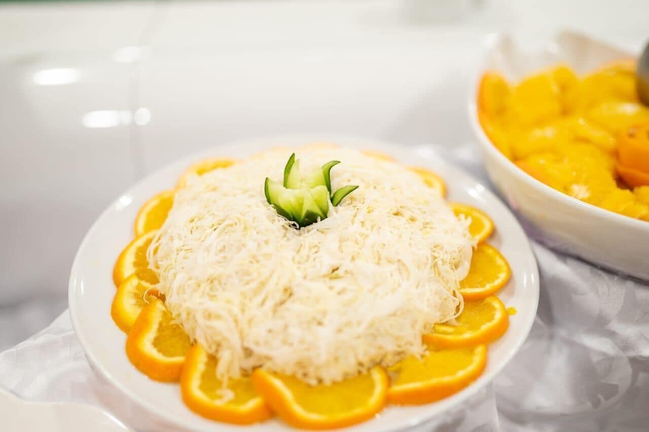 A plate of shredded white food with a green garnish, surrounded by orange slices, on a white tablecloth.