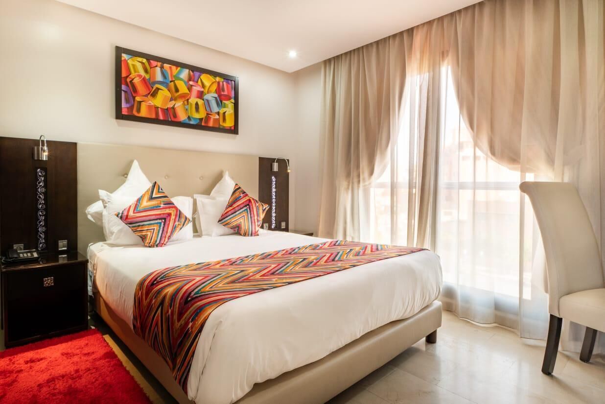 Une chambre d’hôtel avec un grand lit, un tapis rouge, une chaise, un tableau au mur et une fenêtre avec des rideaux.