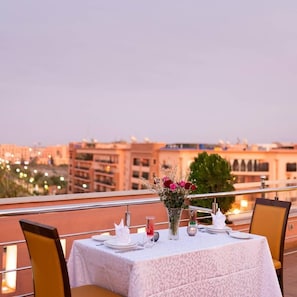 A dining table set for two with a white tablecloth, a vase of flowers, and a cityscape view in the background.