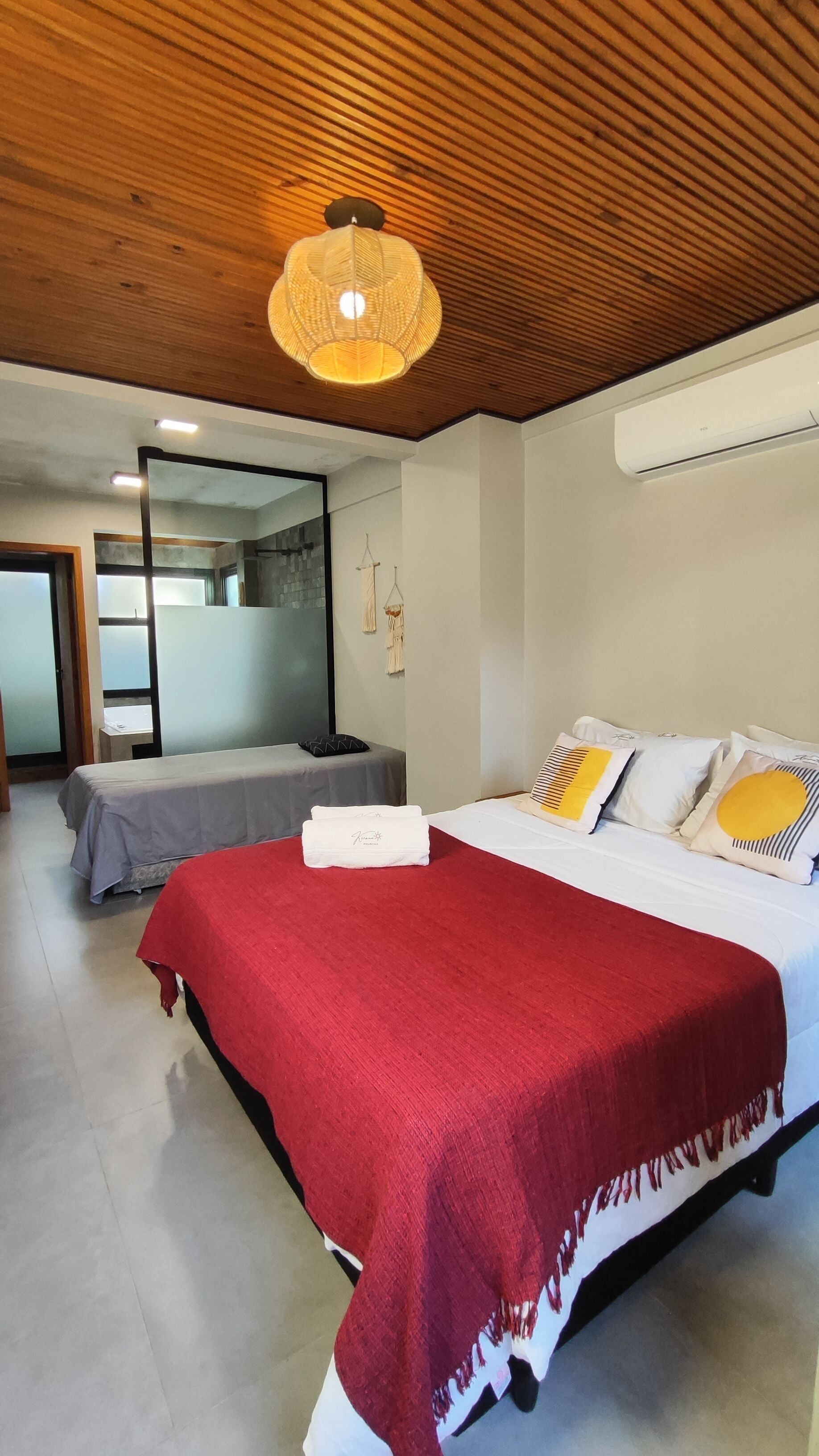 A hotel room with a large bed, a smaller bed, a red blanket, a hanging light fixture, and a wall-mounted air conditioner.