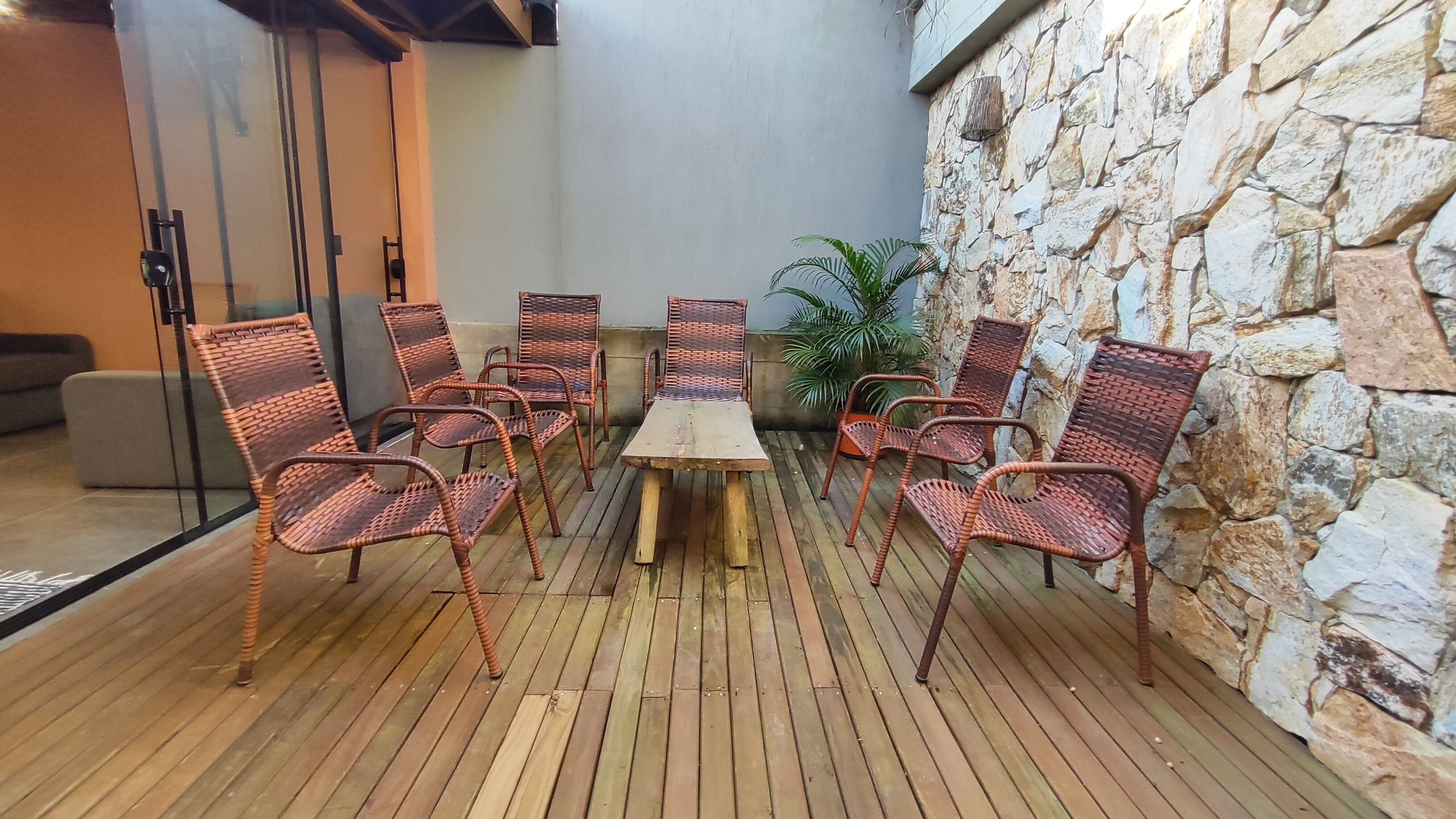 Una terraza de madera con sillas de mimbre y una mesa, un muro de piedra y una puerta de cristal.