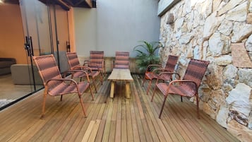 Una terraza de madera con sillas de mimbre y una mesa, un muro de piedra y una puerta de cristal.