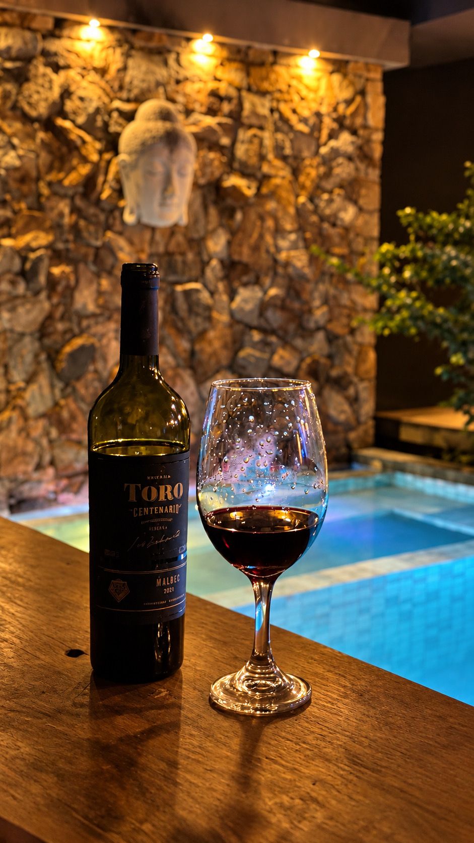 Una ampolla de vi Toro Centenario i un got de vi vermell sobre una taula de fusta, amb una paret de pedra i una piscina al fons.