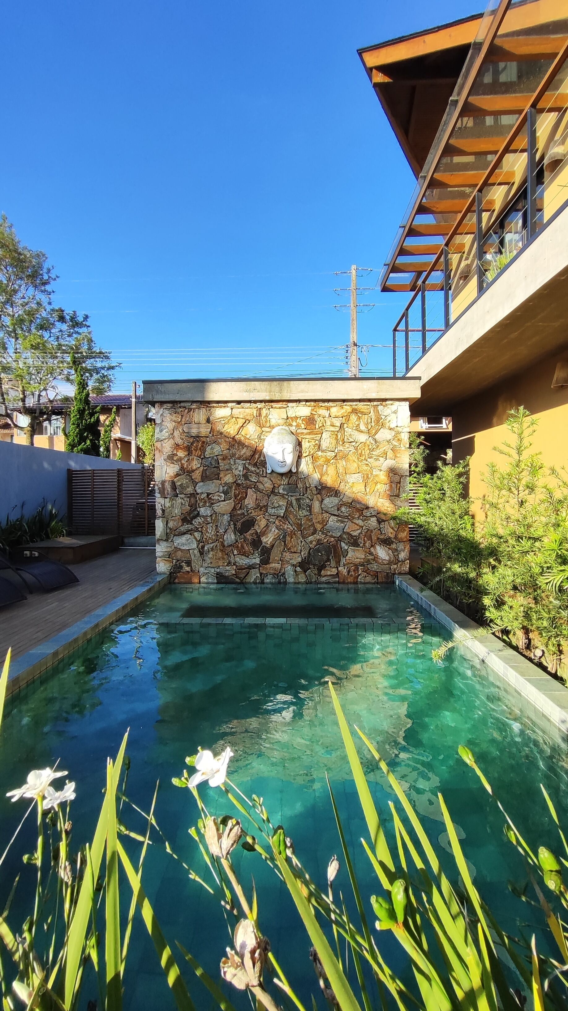 Una piscina con muro de piedra y estatua de Buda, rodeada de follaje y un edificio moderno.