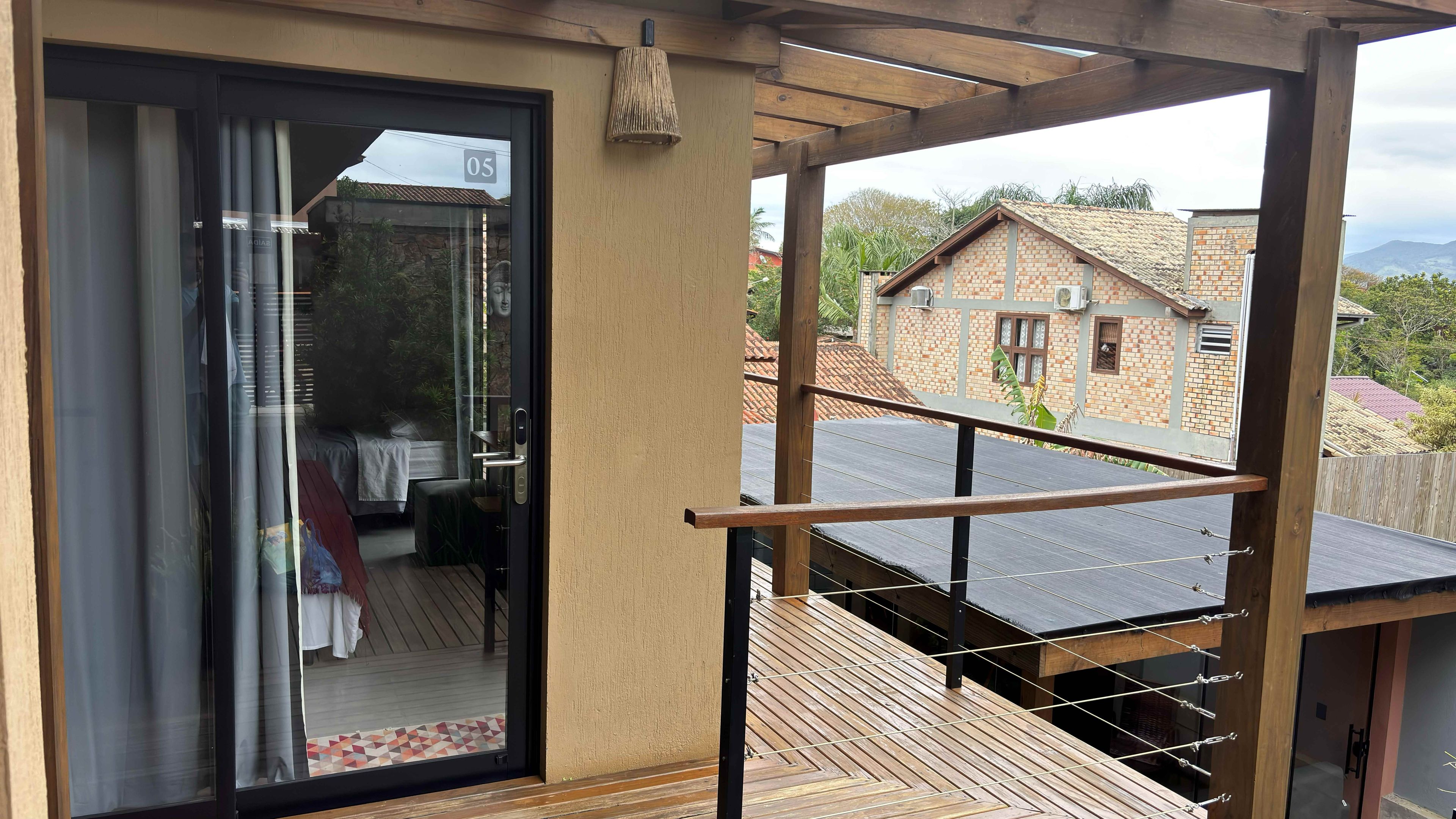A balcony with a wooden railing, a glass door leading to a room with a bed and a table, and a view of a brick house in the background.