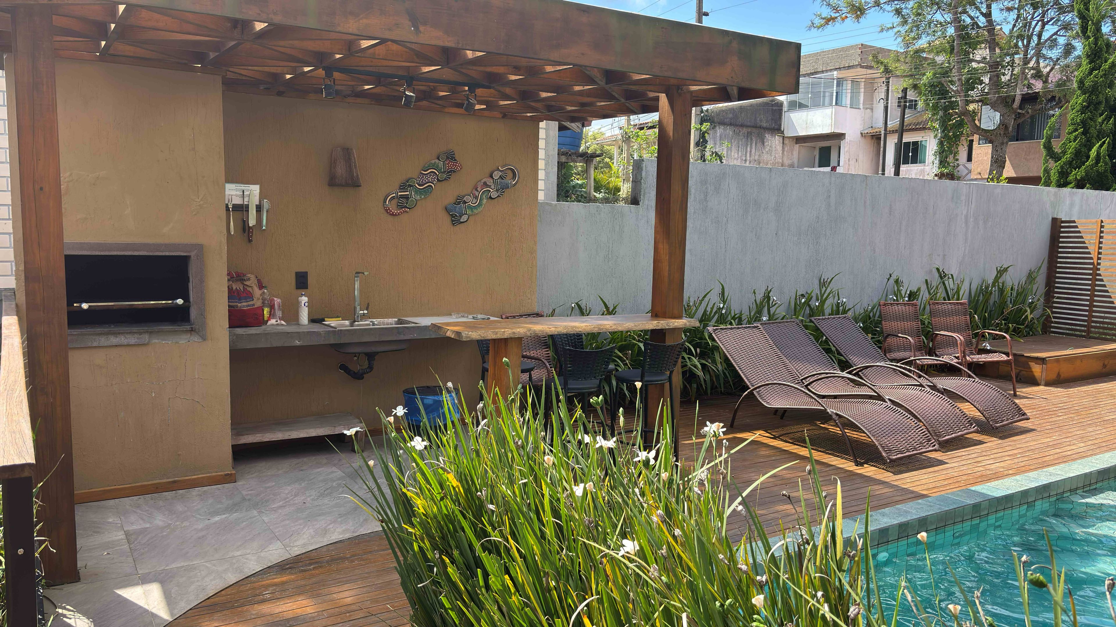 An outdoor kitchen area with a built-in grill, a sink, and a window. There are lounge chairs and a pool in the background.