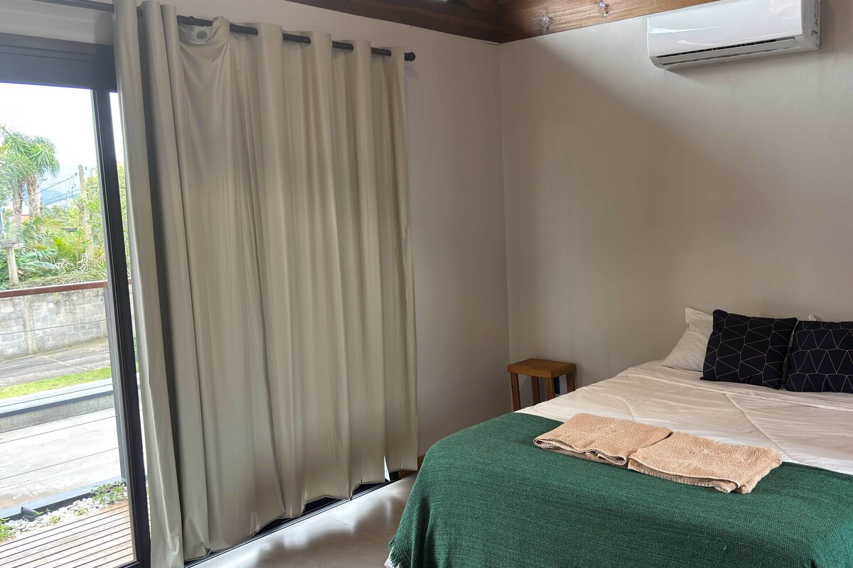 A bedroom with a bed, a small wooden stool, a green blanket, and an air conditioning unit.