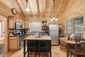 A wooden cabin interior with a kitchen, dining area, and a clock on the wall. - Little Rock Creek Cabin 1BDR (Cherry Log)