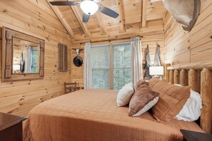 A wooden cabin room with a bed, a ceiling fan, and a large window with blinds. - Little Rock Creek Cabin 1BDR (Cherry Log)