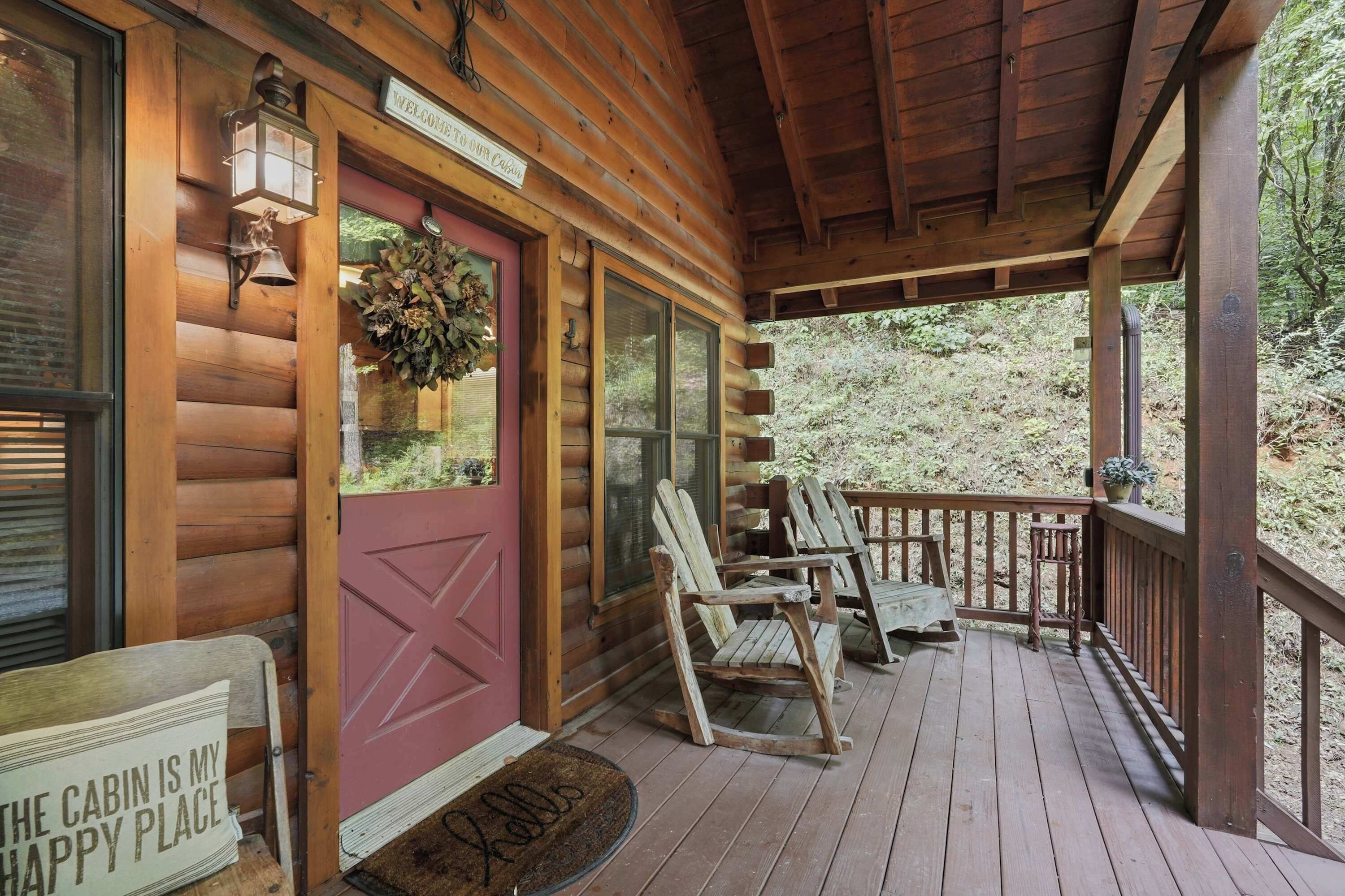 A wooden cabin with a red door, a welcome sign, a porch with wooden chairs, and a sign stating 'The cabin is my happy place.'