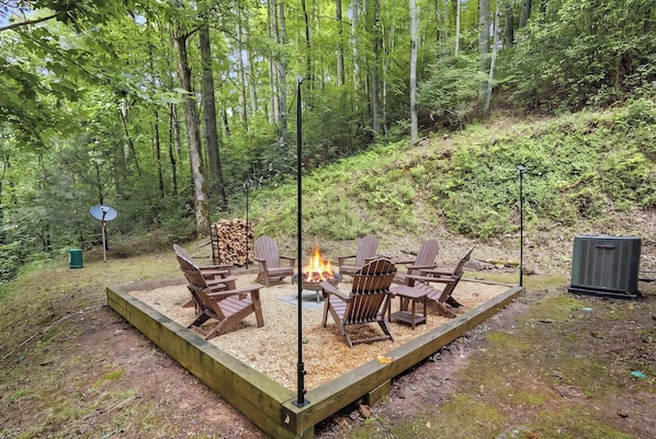 An outdoor seating area with Adirondack chairs around a fire pit, surrounded by a wooden deck and a forested hillside. - Little Rock Creek Cabin 1BDR (Cherry Log)