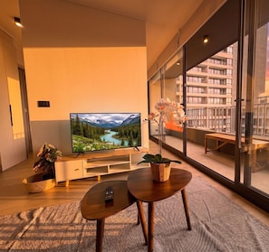 Una sala de estar moderna con un televisor de pantalla plana grande, una mesa de centro de madera, una planta en maceta y vistas de un edificio a través de amplios ventanales.