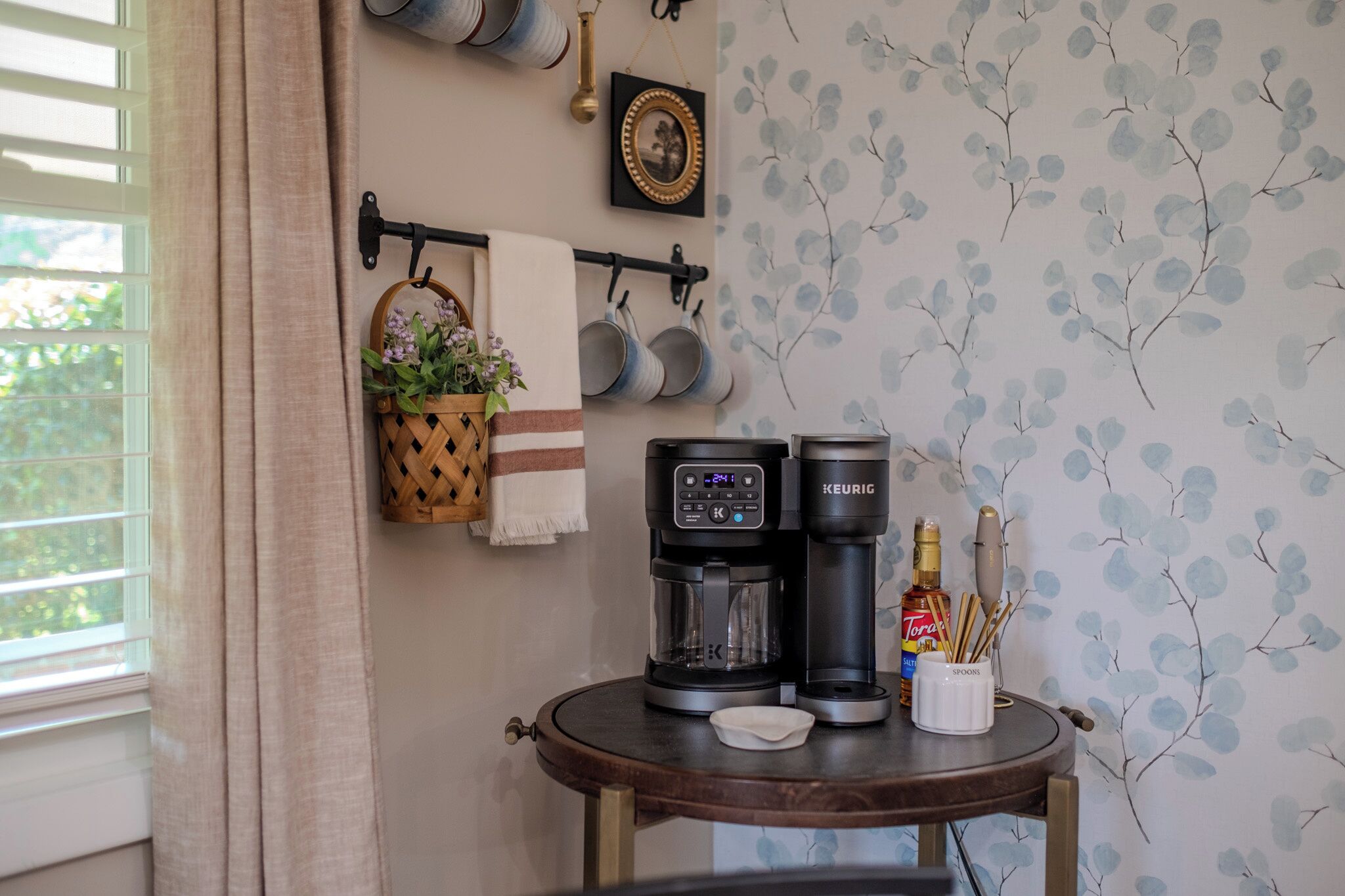 Un área de cocina con una cafetera, una botella de licor y un soporte para cucharas sobre una mesa redonda.
