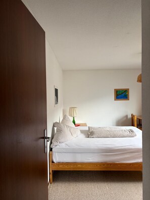 A bedroom with a wooden bed, white bedding, a green lamp, and a painting on the wall.