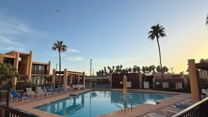 En swimmingpool med liggestole og palmer, omgivet af en murstensplads og et metalhegn.