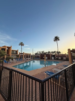 Ein Pool mit Liegestühlen und Palmen, umgeben von einer Ziegelterrasse und einem Metallzaun.