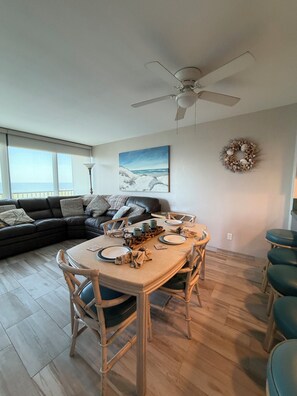 A modern living room with a dining area, a ceiling fan, a painting, and a decorative wreath on the wall. - Breathtaking Ocean View, 3 Cozy bedroom/2 bath Condo. Swimming pool/Tennis court (South Padre Island)