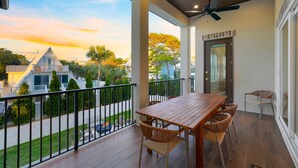 Un balcon avec une table en bois et des chaises en osier, donnant sur un quartier résidentiel avec des maisons et des arbres.