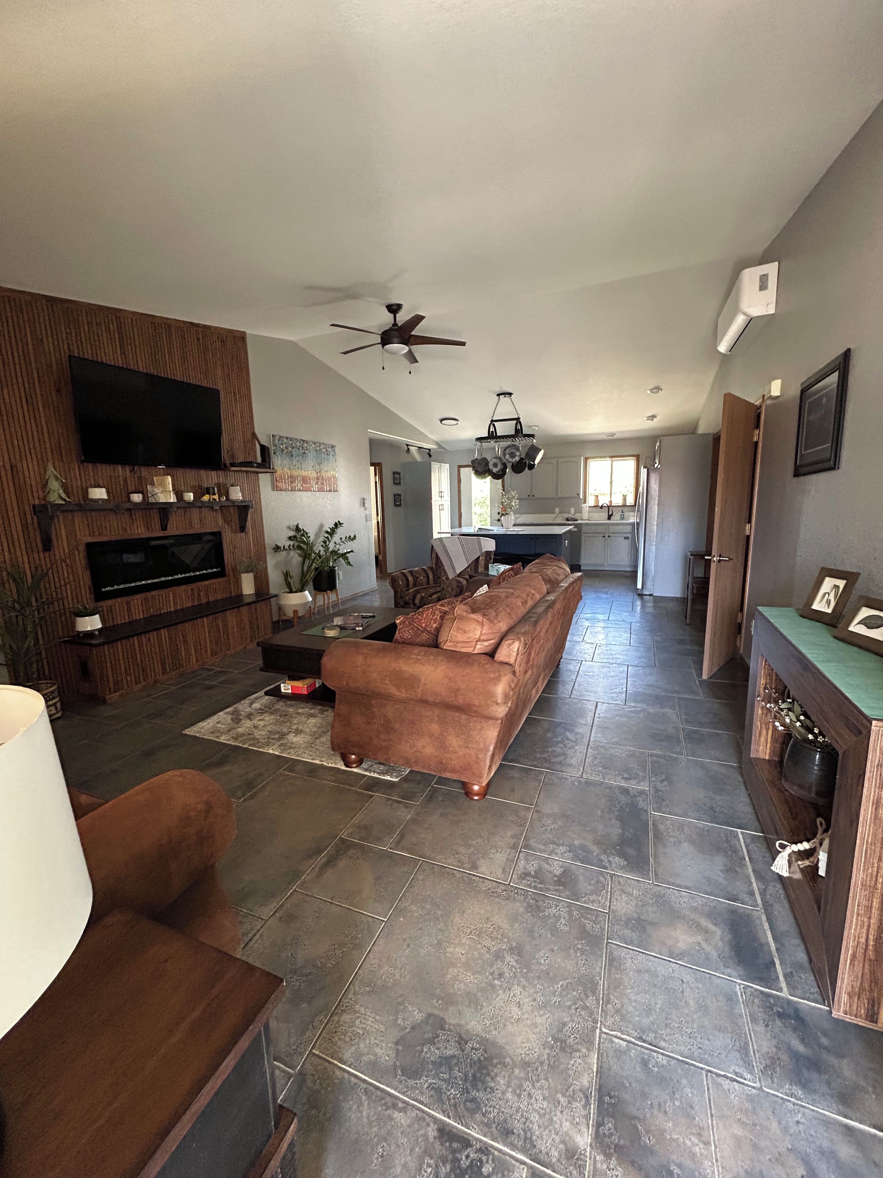 A spacious living area with a large sofa, a flat-screen TV mounted on a wooden wall, a ceiling fan, and a kitchen in the background.