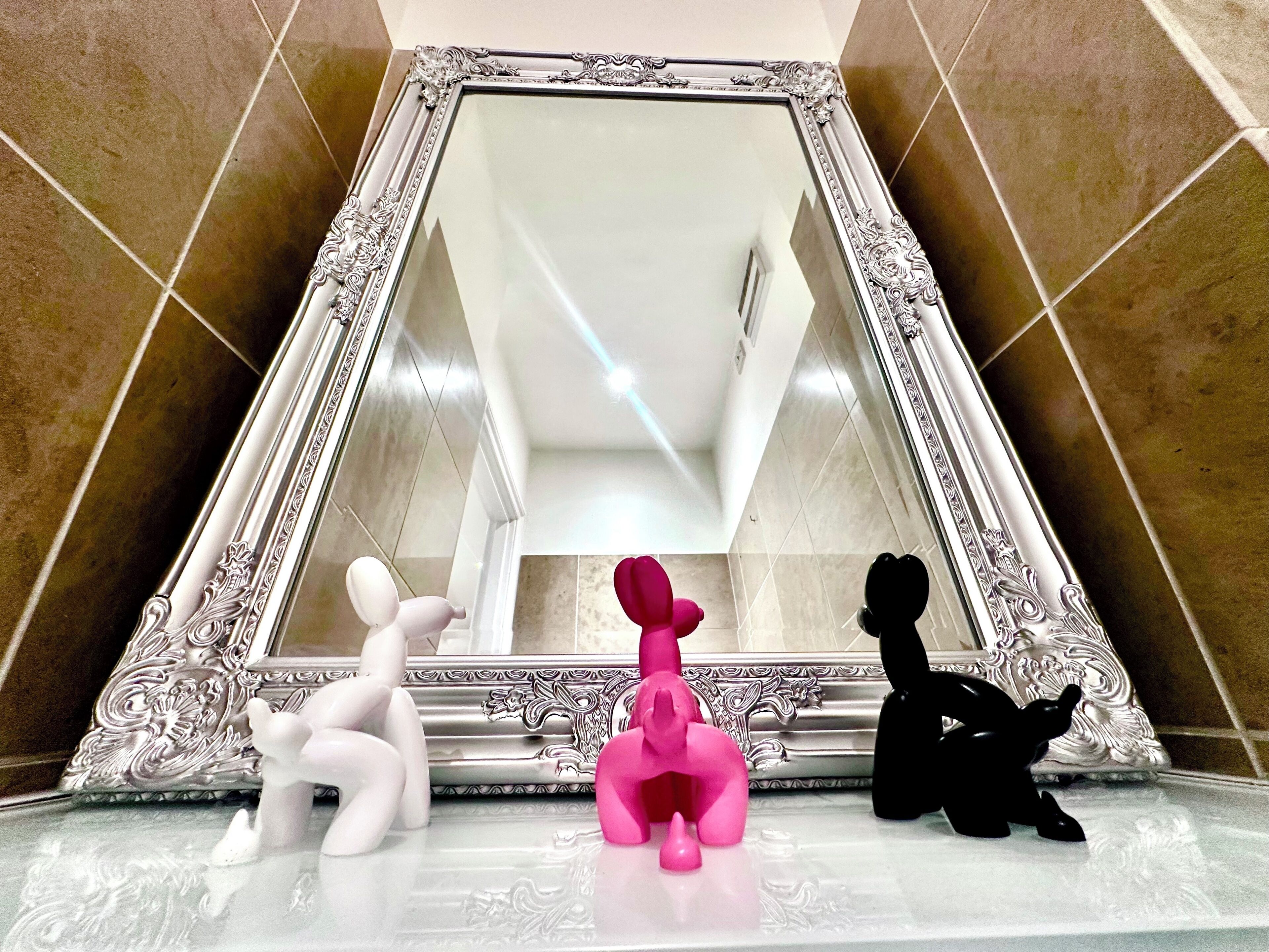 Une salle de bain avec un grand miroir décoratif, une sculpture blanche et rose, et une sculpture noire posée sur le sol.