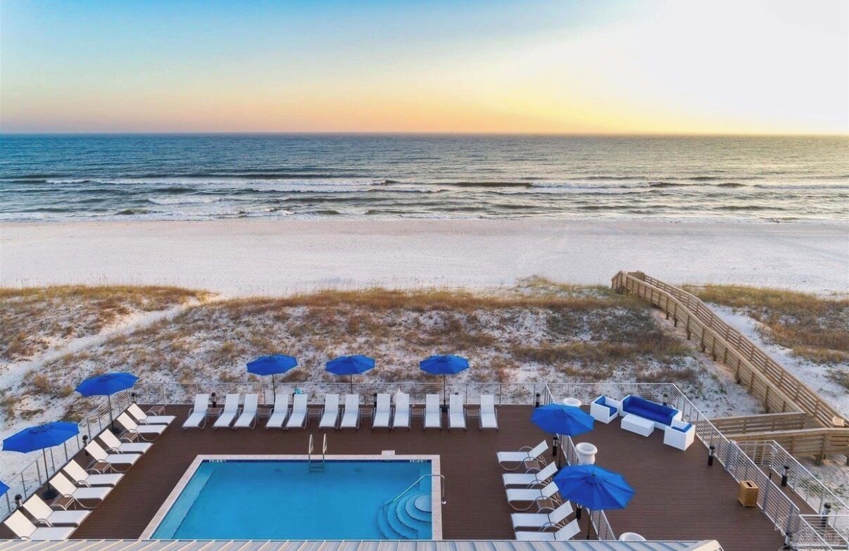 A pool area with lounge chairs and umbrellas, overlooking a beach with a wooden deck and a wooden fence.