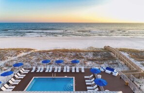 A pool area with lounge chairs and umbrellas, overlooking a beach with a wooden deck and a wooden fence.
