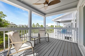 A covered porch with two chairs and a ceiling fan, overlooking a pool and greenery.