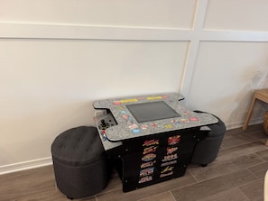 A small, portable gaming table with a joystick, surrounded by two ottomans, against a plain wall.