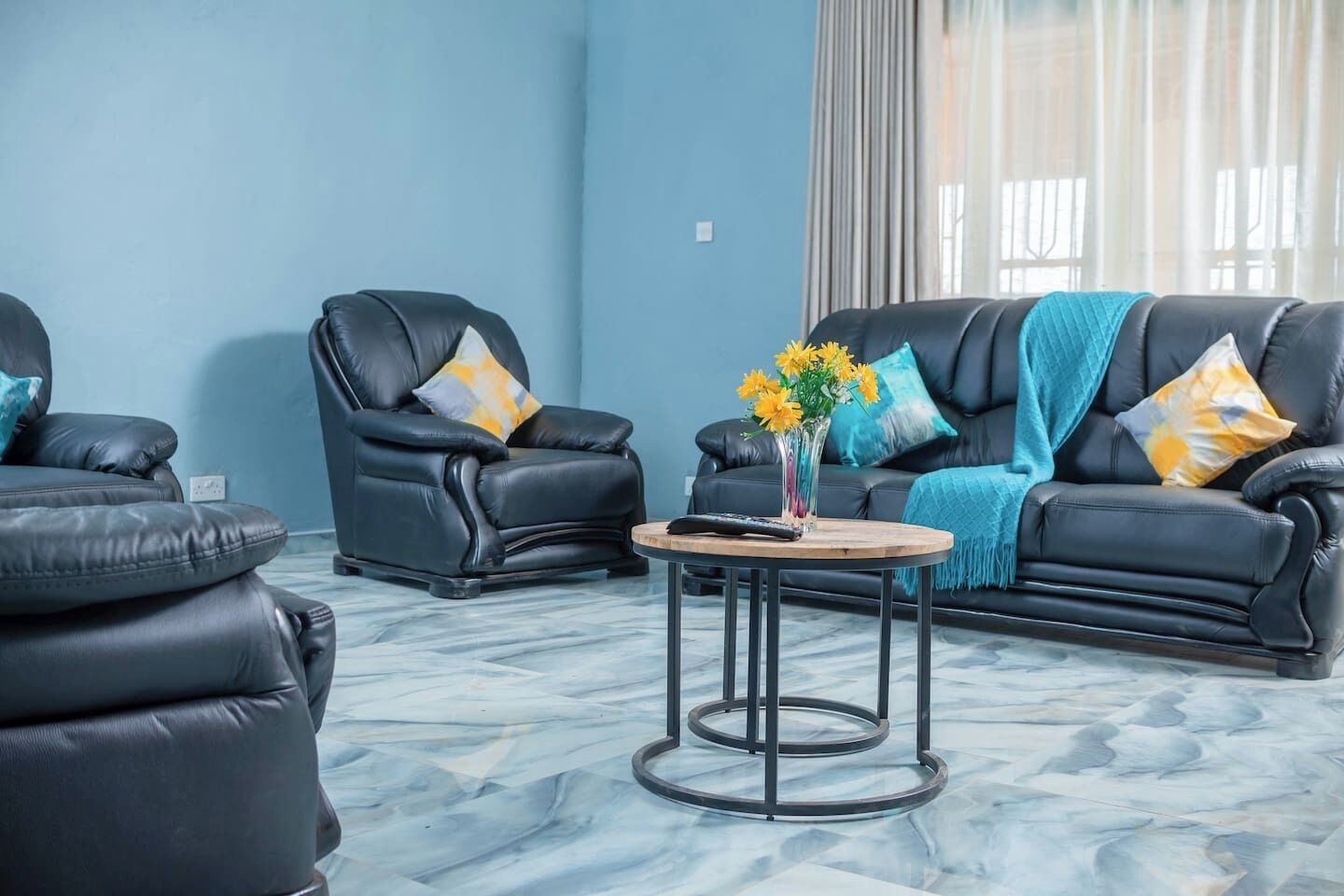 Un salon moderne avec un mur bleu, deux canapés en cuir noir ornés de coussins à motifs, une table basse en bois sur laquelle est posé un vase de fleurs jaunes, et une grande fenêtre avec des rideaux.