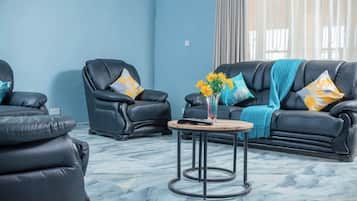 A modern living room with a blue wall, two black leather sofas with patterned cushions, a wooden coffee table with a vase of yellow flowers, and a large window with curtains.