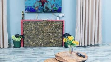 A room with a flat-screen TV mounted on the wall, a round wooden table with a vase of flowers, and a patterned cabinet below the TV.